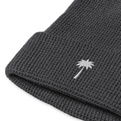 A close-up of a dark gray Mambí knitted beanie with a folded edge, featuring a small white embroidered palm tree on the cuff.
