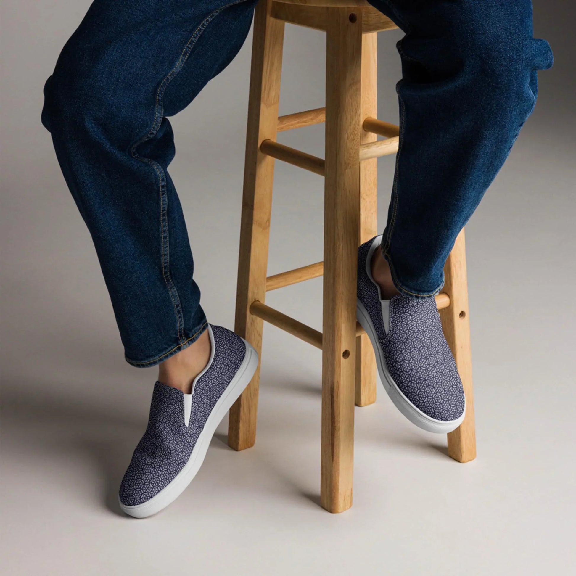 A person sits on a light wooden stool in blue jeans and Printfuls Digno Handmade Slip-On Canvas Shoes for men, with only the lower half visible, making a subtle fashion statement against a plain background.