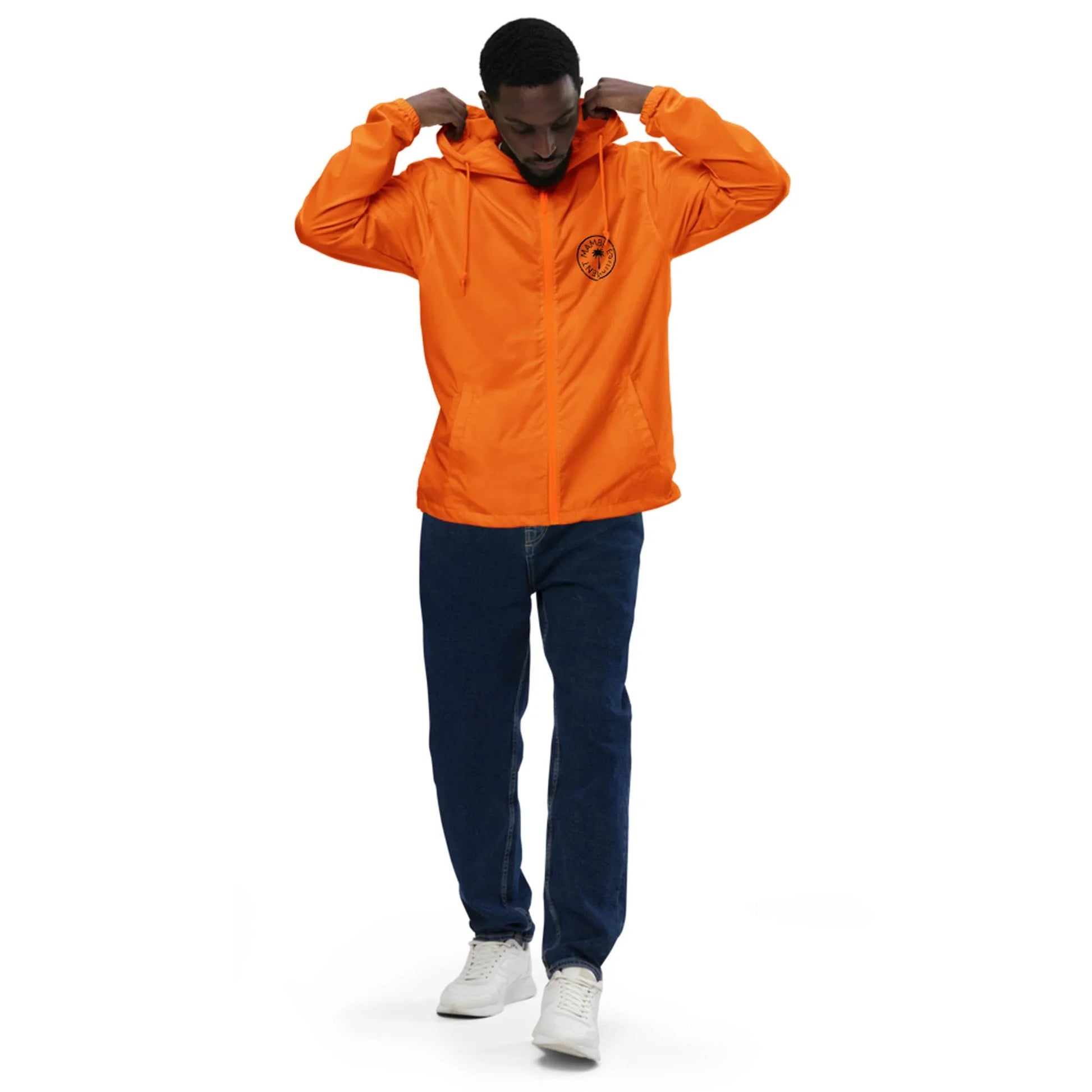 A man adjusts the hood of his bright orange Mambí jacket, featuring a small black emblem on the chest. He is wearing dark blue jeans and white sneakers, standing against a plain white background.