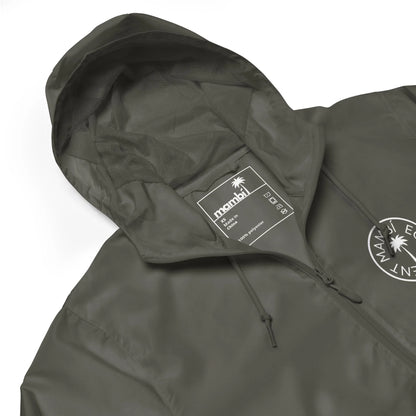 A close-up of an olive green hooded jacket with a drawstring hood. The label inside reads “manahil” with size and care details. A circular Mambí logo featuring text and a palm tree decorates the chest.