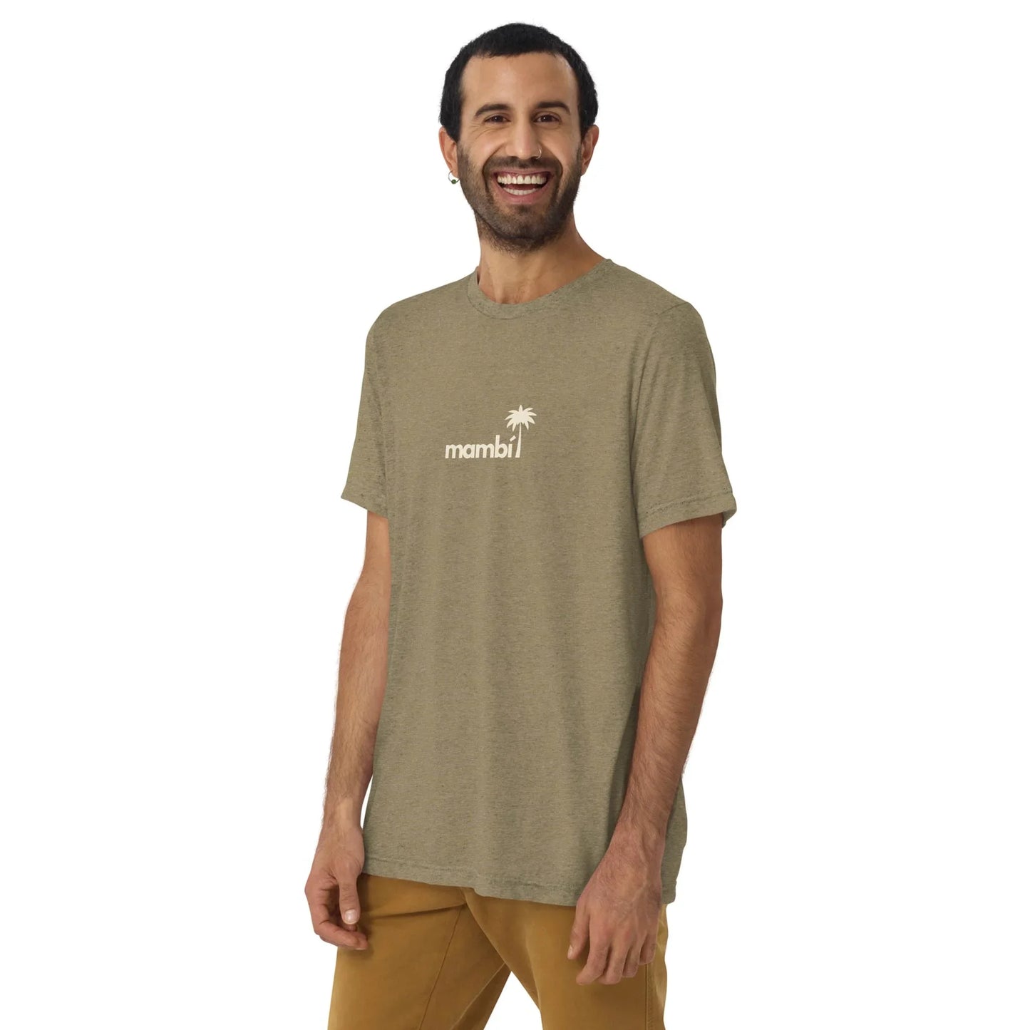 A man with short dark hair and a beard smiles in a Printful El Comemierda Tri-Blend Graphic Tee (unisex) featuring a palm tree graphic, light olive green color, and tan pants, standing against a plain white background.