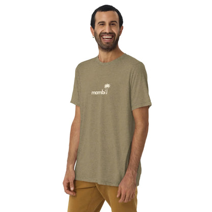 A man with short dark hair and a beard smiles in a Printful El Comemierda Tri-Blend Graphic Tee (unisex) featuring a palm tree graphic, light olive green color, and tan pants, standing against a plain white background.