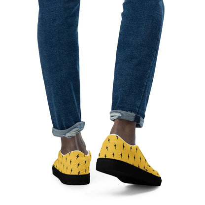 A person is seen from behind, wearing rolled-up blue jeans and Printfuls Electra Handmade Slip-On Canvas Shoes for men, featuring a bold black lightning bolt design—an eye-catching fashion statement against a white background.