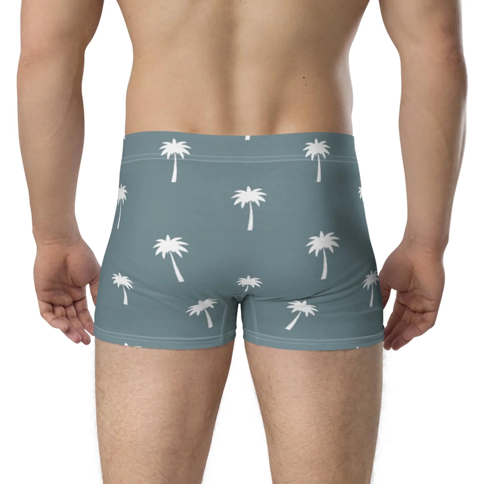 A man, shown from the back, wears Printfuls Emerson Boxer Brief for men in light blue with a white palm tree pattern. The boxer brief features a no-back-seam design and seamless fit for comfort, set against a plain neutral background.
