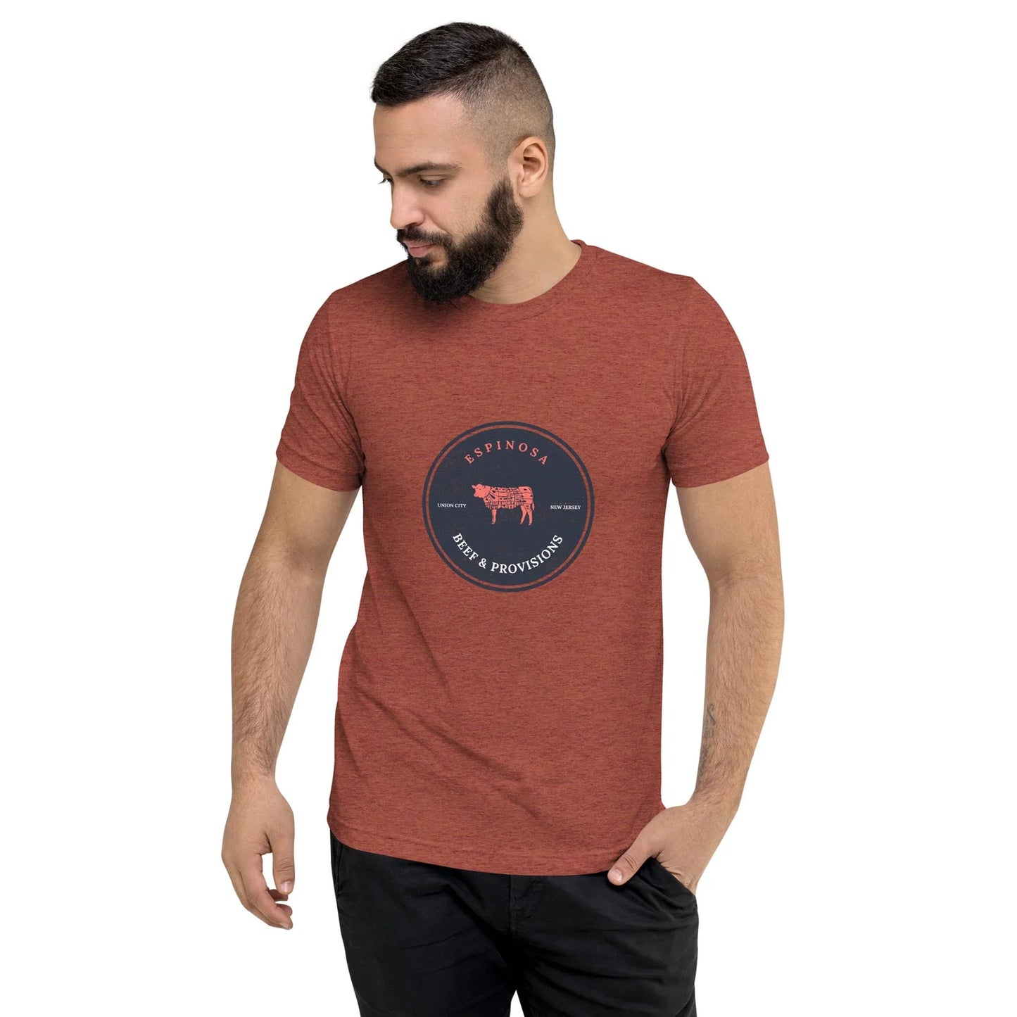 A man with a beard wears a red heather Mambí t-shirt featuring a circular Espinosa Iberian Provisions logo with a pig illustration in the center, standing with one hand in his pocket and looking down to his left.