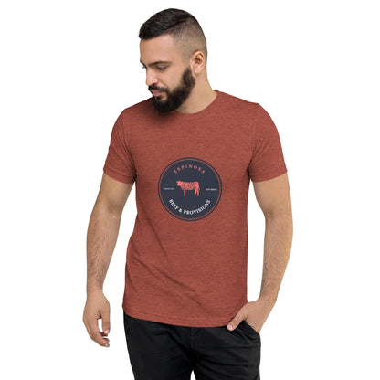 A man with a beard wears a red heather Mambí t-shirt featuring a circular Espinosa Iberian Provisions logo with a pig illustration in the center, standing with one hand in his pocket and looking down to his left.