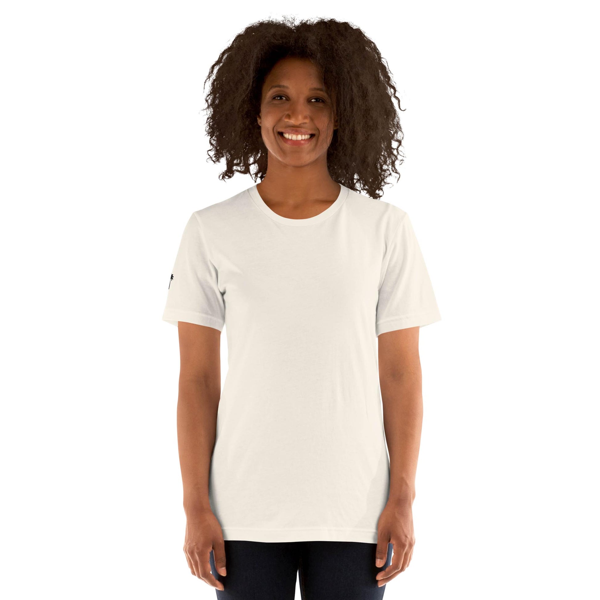 A woman with curly hair smiles while standing and wearing a plain white Mambí T-shirt and dark pants against a white background.