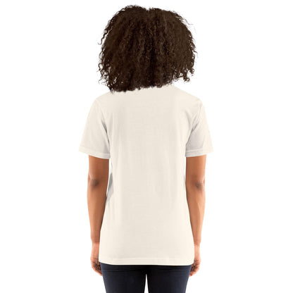 A person with curly hair is standing with their back to the camera, wearing a plain white Mambí short-sleeve t-shirt and dark jeans against a white background.