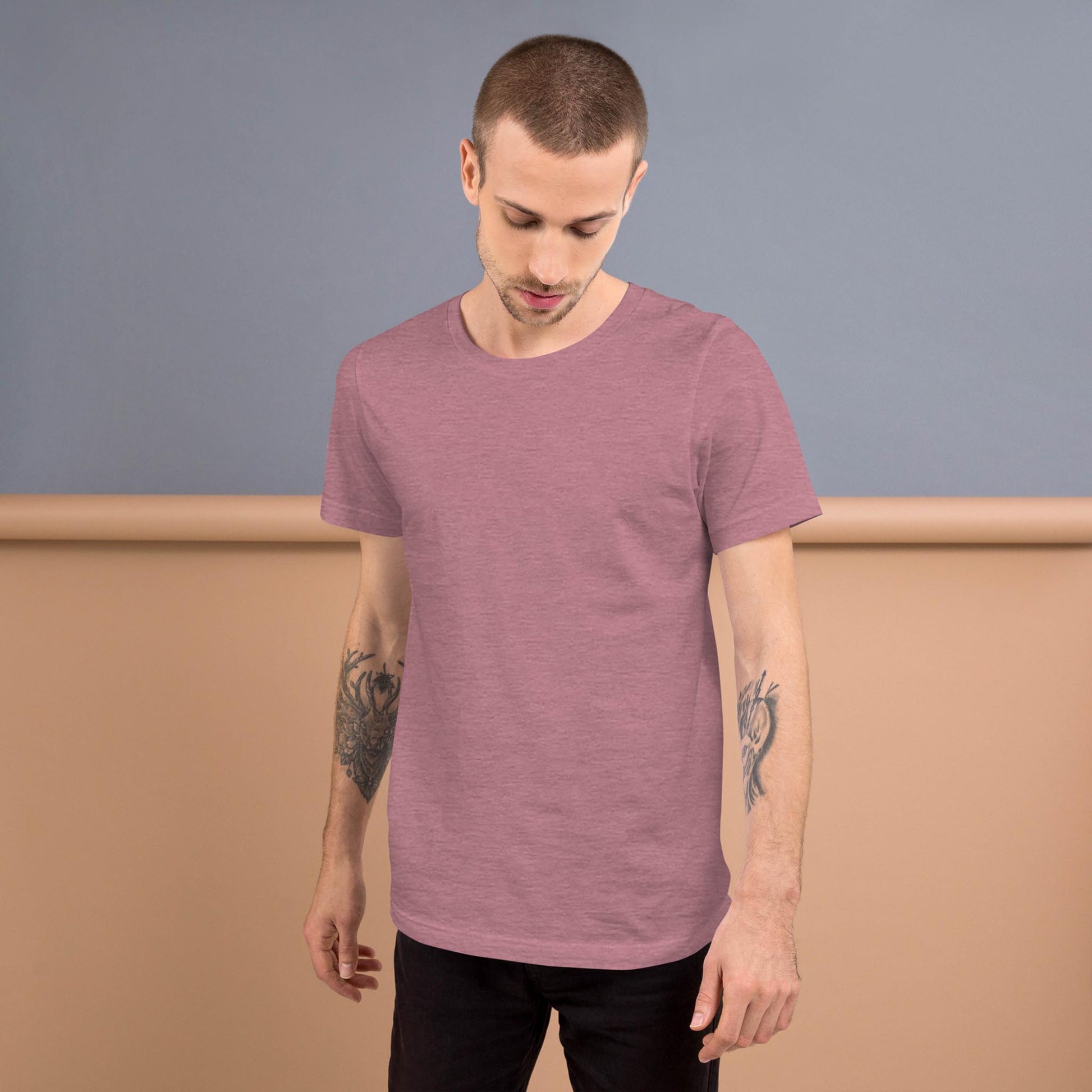 A man with short hair and arm tattoos is wearing a muted mauve Mambí t-shirt and black pants, standing against a two-tone beige and gray background, looking down with a neutral expression.