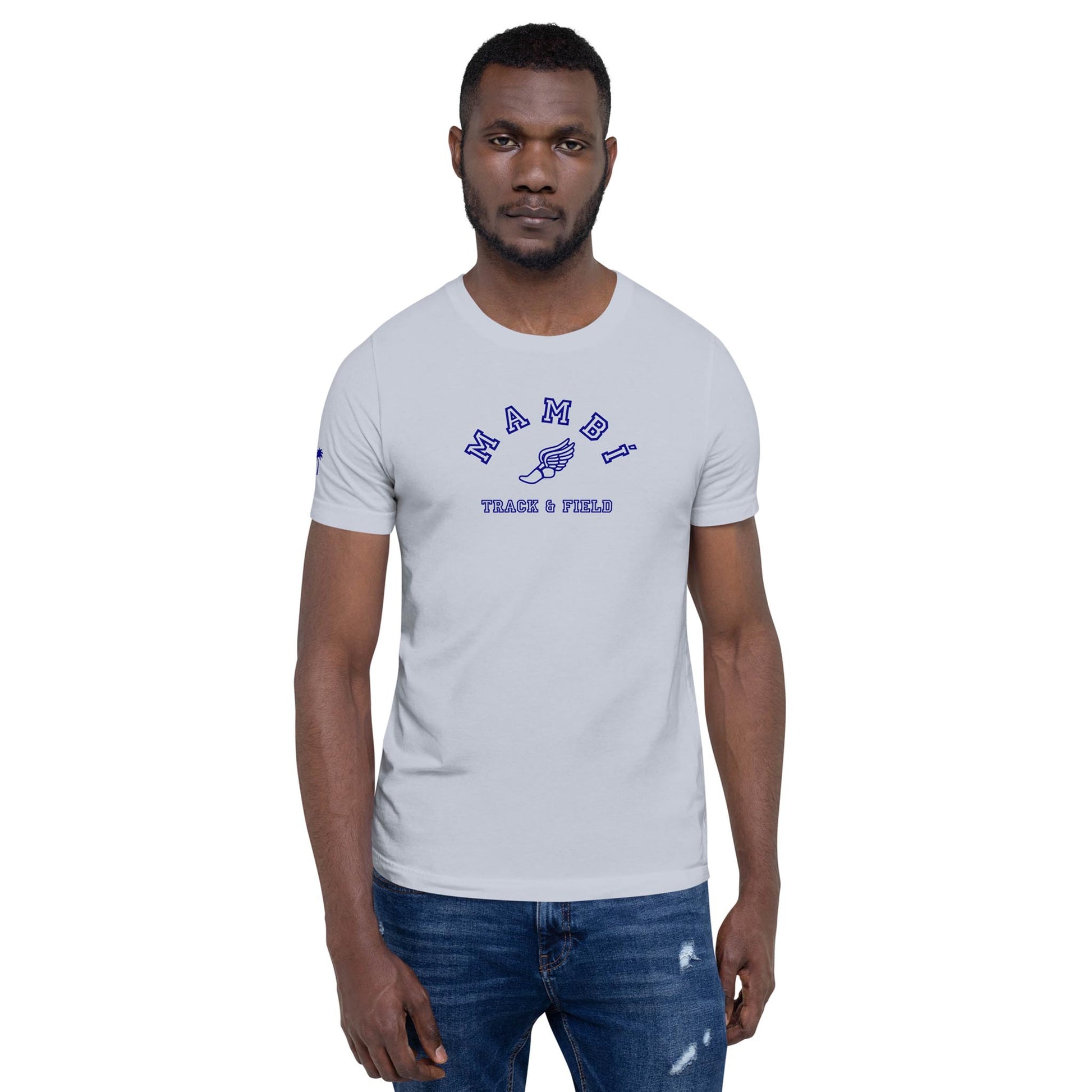 A man with a beard wears a light gray Mambí t-shirt that says NAMIBI TRACK & FIELD in blue text. He stands facing forward with a neutral expression and is also wearing slightly distressed blue jeans.