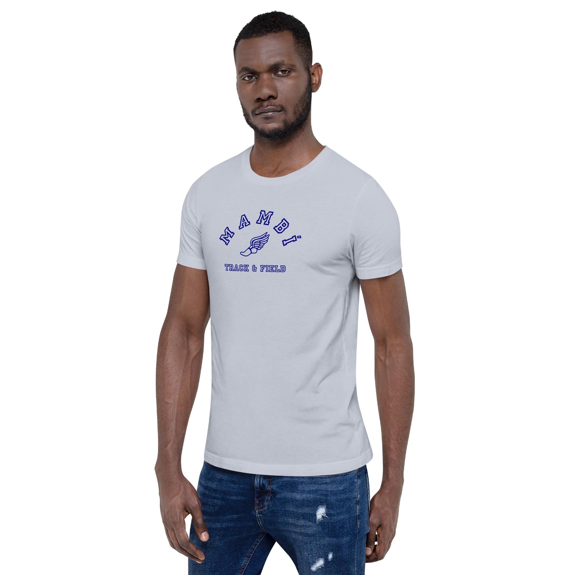 A man in a light gray Mambí t-shirt with blue text reading NAMDI TRACK & FIELD stands facing forward, one hand by his side and the other slightly bent, wearing ripped blue jeans.