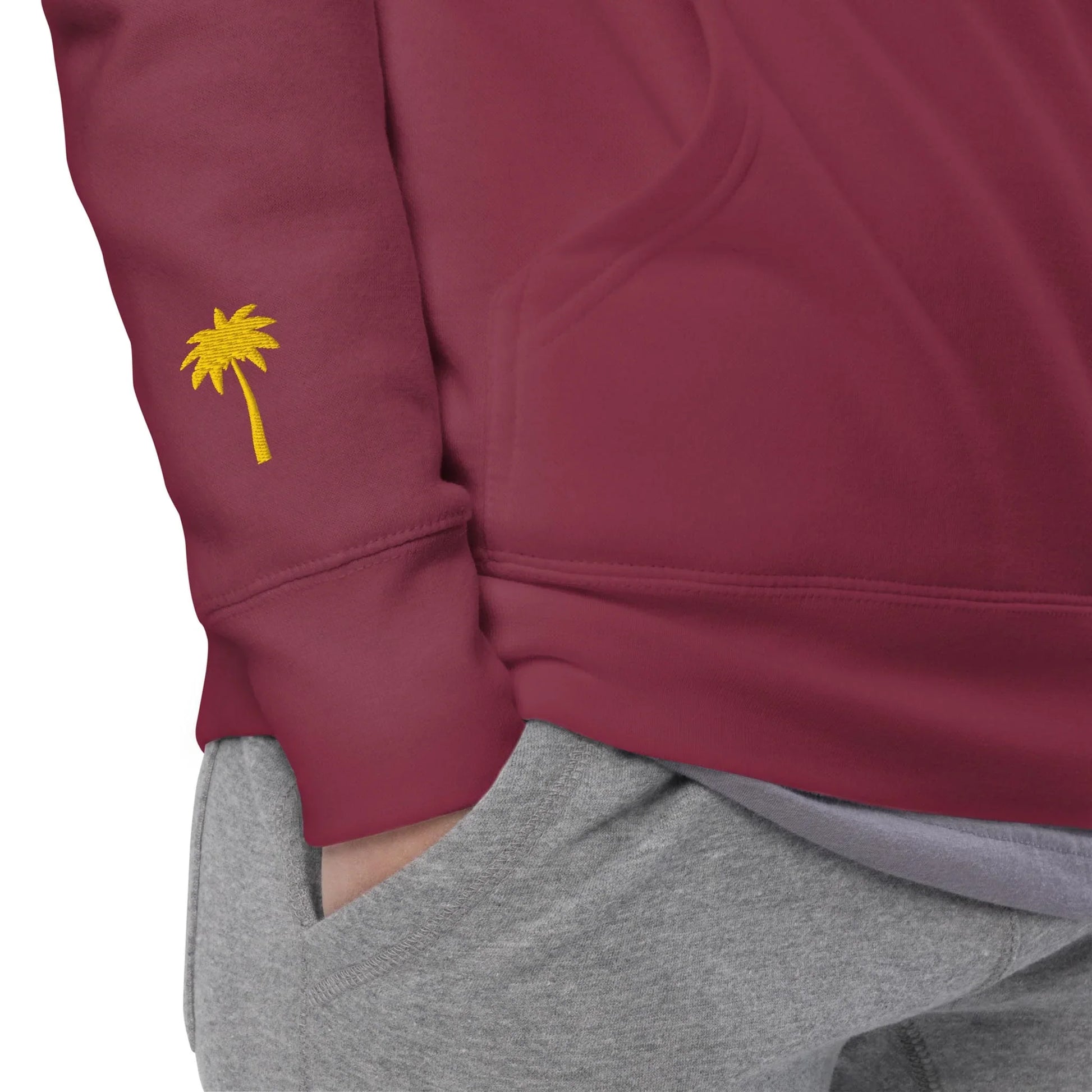 A person wearing a maroon Mambí sweatshirt with a small yellow palm tree embroidered on the sleeve and gray sweatpants, with one hand in their pocket.