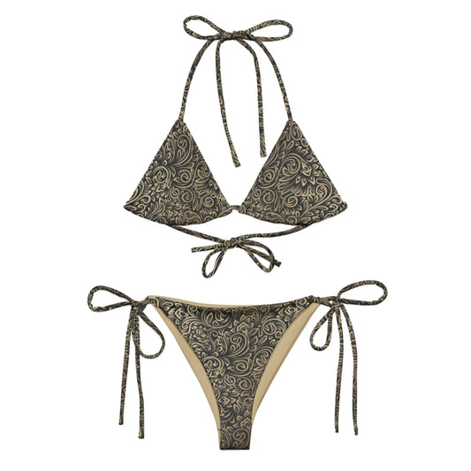 The Flavia String Bikini Swimsuit by Printful features a black and gold swirling floral pattern, triangle cups with ties, side-tie bottoms, recycled polyester fabric, and UPF 50+ sun protection.