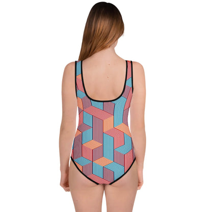 A woman with long brown hair stands facing away, wearing a Mambí one-piece swimsuit adorned with a colorful geometric pattern of interlocking striped shapes in blue, red, orange, and white.