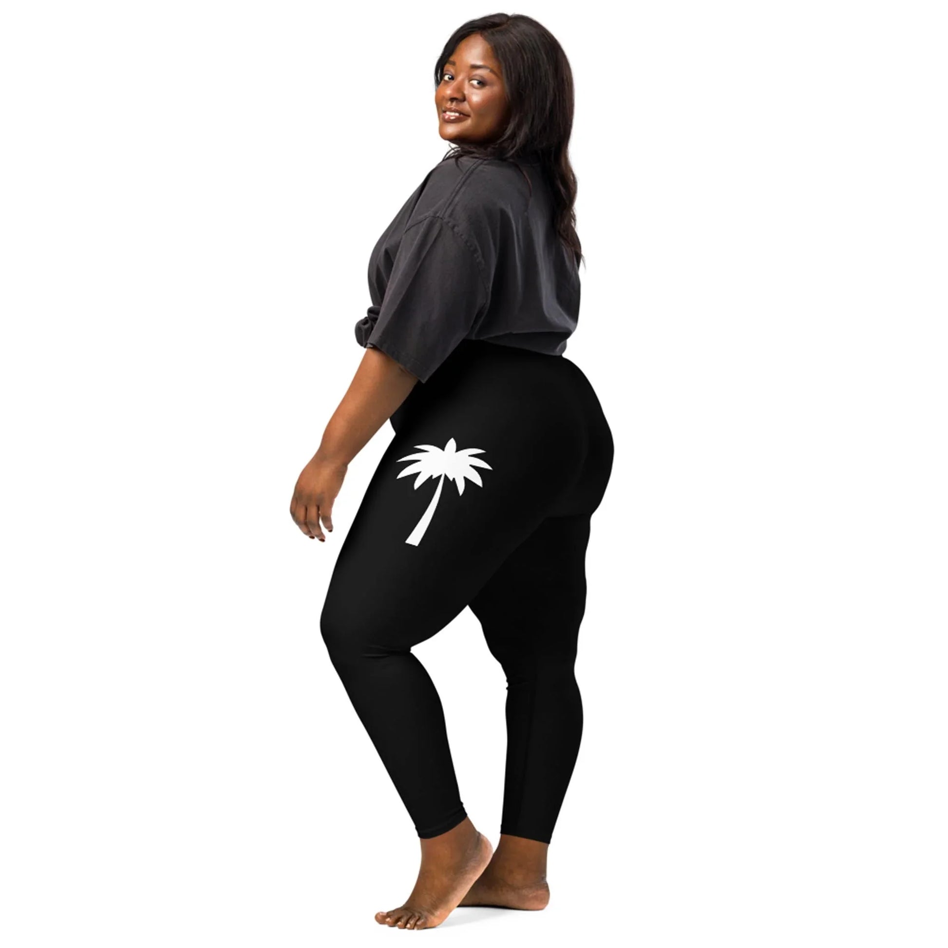 A woman with long dark hair wears a loose black Mambí t-shirt and black leggings featuring a white palm tree graphic on the upper thigh, standing barefoot against a plain white background.