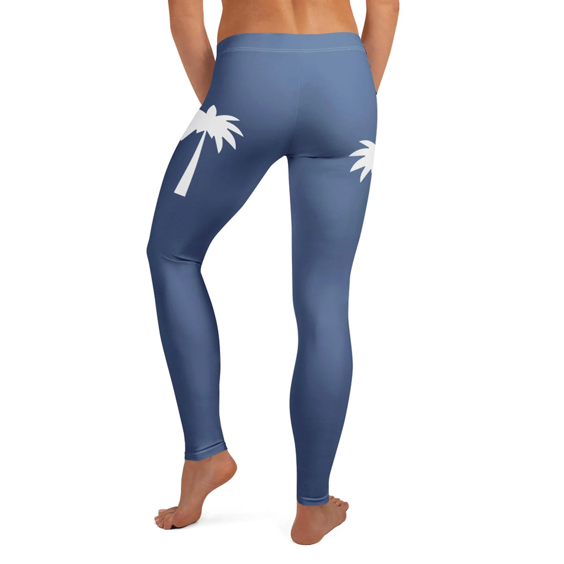 A person wearing high-waisted blue Mambí leggings with white palm tree graphics on the back, standing barefoot and facing away from the camera.