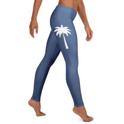 A person wearing high-waisted, blue Mambí leggings with a white palm tree graphic on the upper thigh, standing barefoot and viewed from the side.