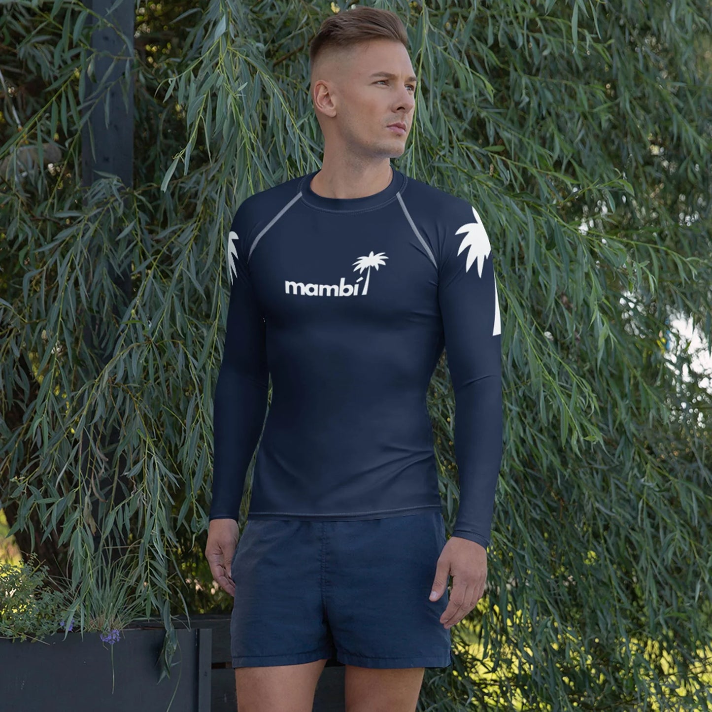 A man stands outdoors by leafy greenery, wearing the Printful Hasselhoff Classic Long-Sleeve Rash Guard for men with UPF 50+ and a white palm tree mambi1 graphic on the chest, paired with navy shorts—ideal for watersports.