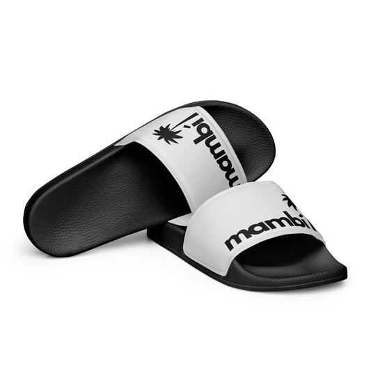 A pair of black and white Mambí slide sandals, one lying flat and the other on its side. The white strap features black text reading mambi alongside a black palm tree graphic.