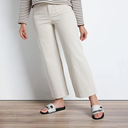 A person wearing a striped long-sleeve shirt, light beige pants, and white Mambí slide sandals with black soles stands on a wooden floor against a white background.