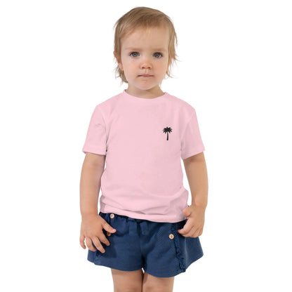 A young child with light hair wears a light pink Mambí t-shirt featuring a small black palm tree graphic on the chest and blue shorts with buttons, standing against a plain white background.