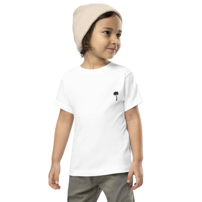 A young child wearing a beige knit beanie, white t-shirt with a small black palm tree graphic on the chest, and khaki pants, smiling and looking to the side against a plain background—styled by Mambí.