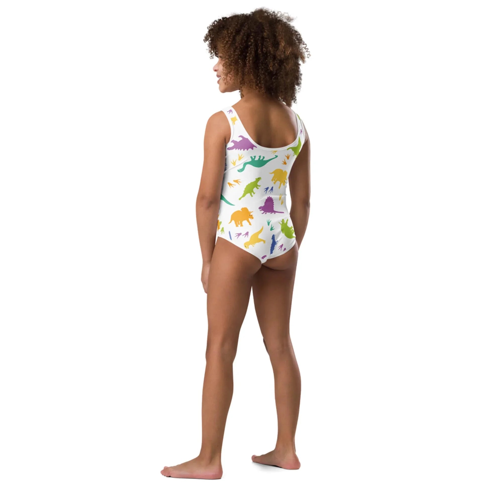 A young girl stands barefoot facing away, wearing a white Mambí one-piece swimsuit with colorful dinosaur patterns. She has curly hair and stands against a plain white background.