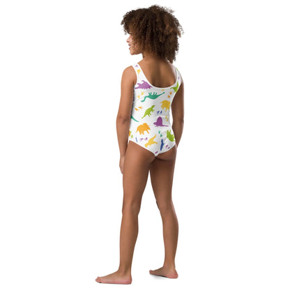 A young girl stands barefoot facing away, wearing a white Mambí one-piece swimsuit with colorful dinosaur patterns. She has curly hair and stands against a plain white background.