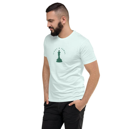 A man with a beard stands, looking to his left, wearing a light-colored Mambí t-shirt with a small chess piece graphic and the words Kings & Pawns on the chest. His hands are in his pockets.