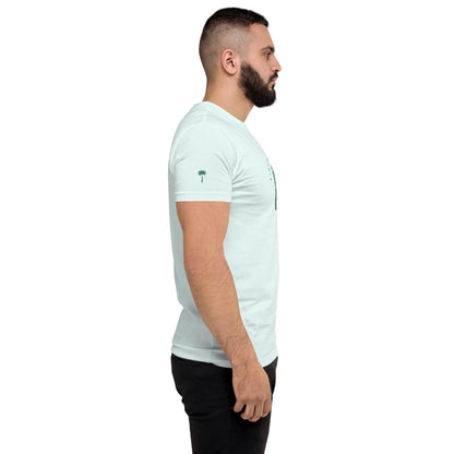 A man with a beard stands in profile, facing left, wearing a light blue Mambí T-shirt and black pants against a plain white background.