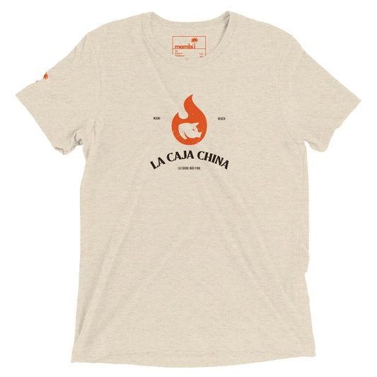 Beige t-shirt featuring a red flame graphic above a pig silhouette and La Caja China printed below, with Mambí-inspired details in small text around the graphic. A logo is visible inside the collar.