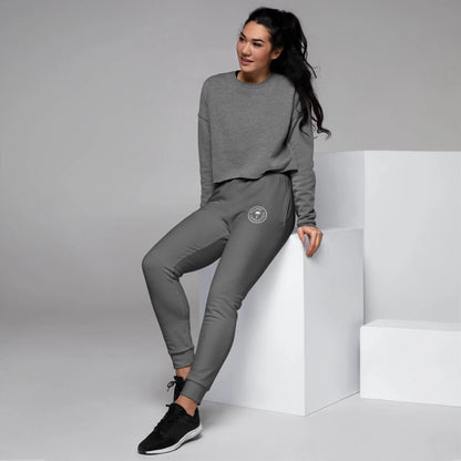 A woman with long dark hair, wearing a gray Mambí sweatshirt and gray joggers, sits on a white block in a minimal studio. She is smiling and dressed in black sneakers.