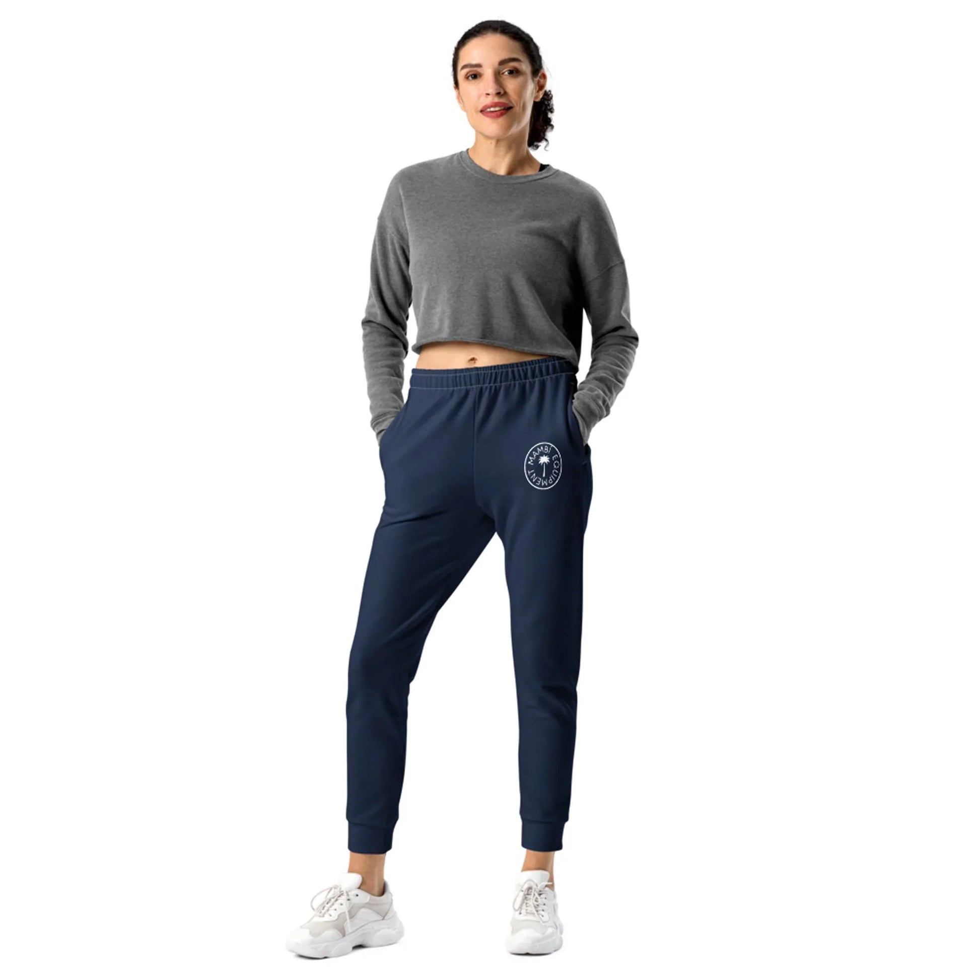 A woman stands against a white background wearing a gray Mambí long-sleeve crop top, navy jogger pants with a circular white logo on one leg, and white sneakers, her hands in her pockets as she smiles.