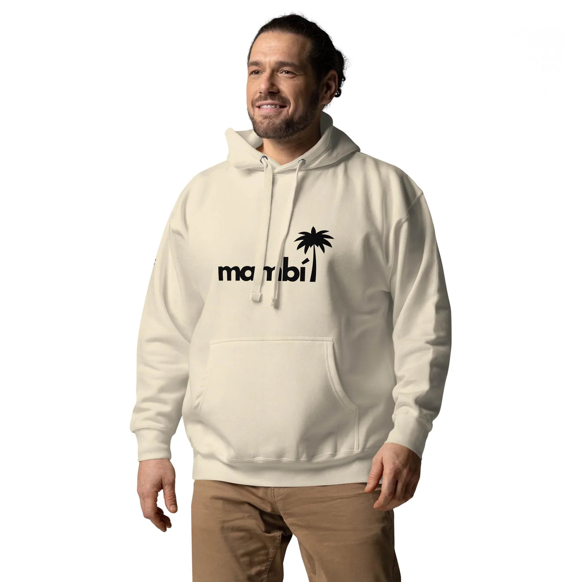 A man wearing a light-colored Mambí hoodie with a miami logo and palm tree design stands smiling against a plain white background. He has dark hair tied back and is also wearing tan pants.