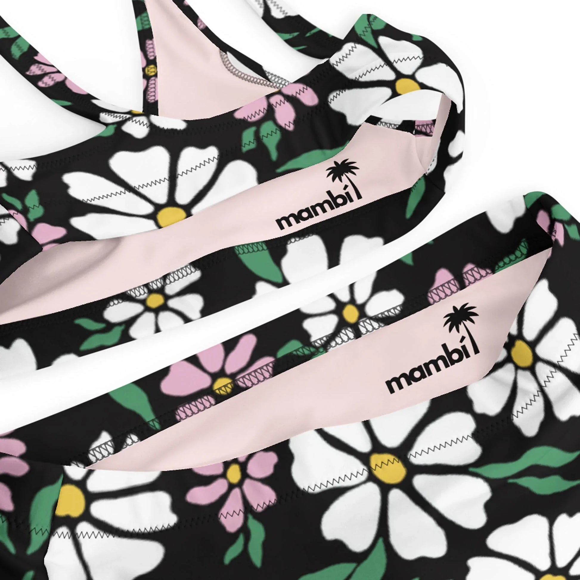 Close-up of a floral-patterned Mambí bikini featuring white, pink, and yellow flowers on a black background. The Mambí name with a palm tree icon is printed on the fabric.