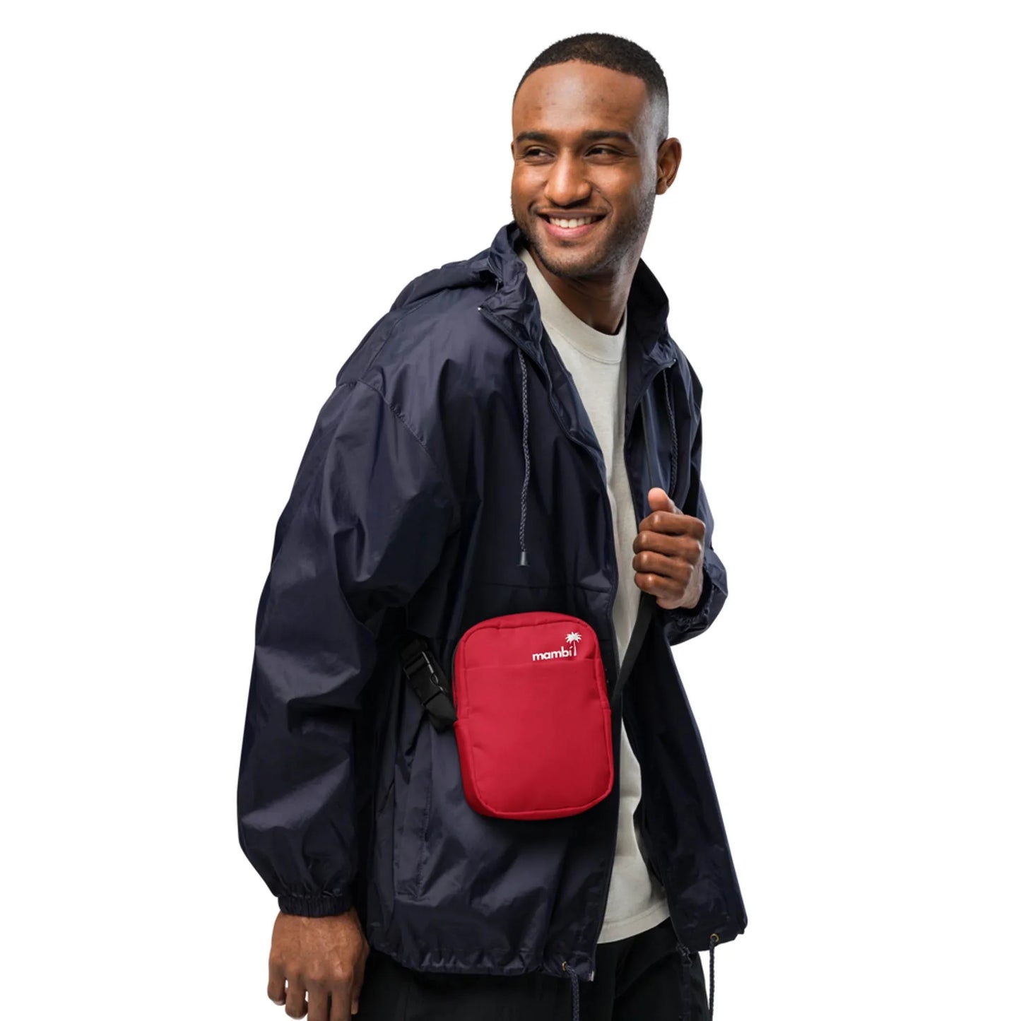 A man in a navy blue Mambí windbreaker and white shirt smiles while standing. He has a small red crossbody bag with a white logo across his chest. The background is plain white.