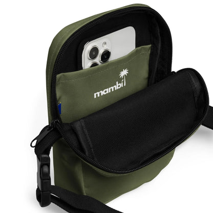 An olive green Mambí crossbody bag with a front pocket, partially unzipped to reveal a smartphone inside. The word mambi and a palm tree logo are printed in white above the pocket.