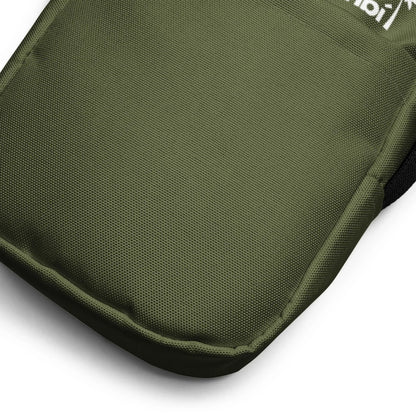 Close-up view of the bottom corner of an olive green Mambí fabric backpack, showing its textured material and reinforced stitching. Part of a white logo is visible at the top edge of the image.