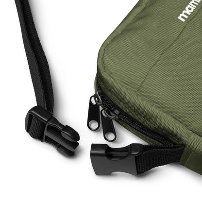 Close-up of a green Mambí fabric bag with black zippers and a black adjustable strap, featuring a plastic buckle clasp. The strap and buckle are unfastened, lying on a white surface.