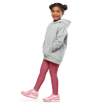 A smiling young girl with curly hair in pigtails wears a gray Mambí hoodie, pink leggings, and pink sneakers. She stands with one foot forward against a white background.