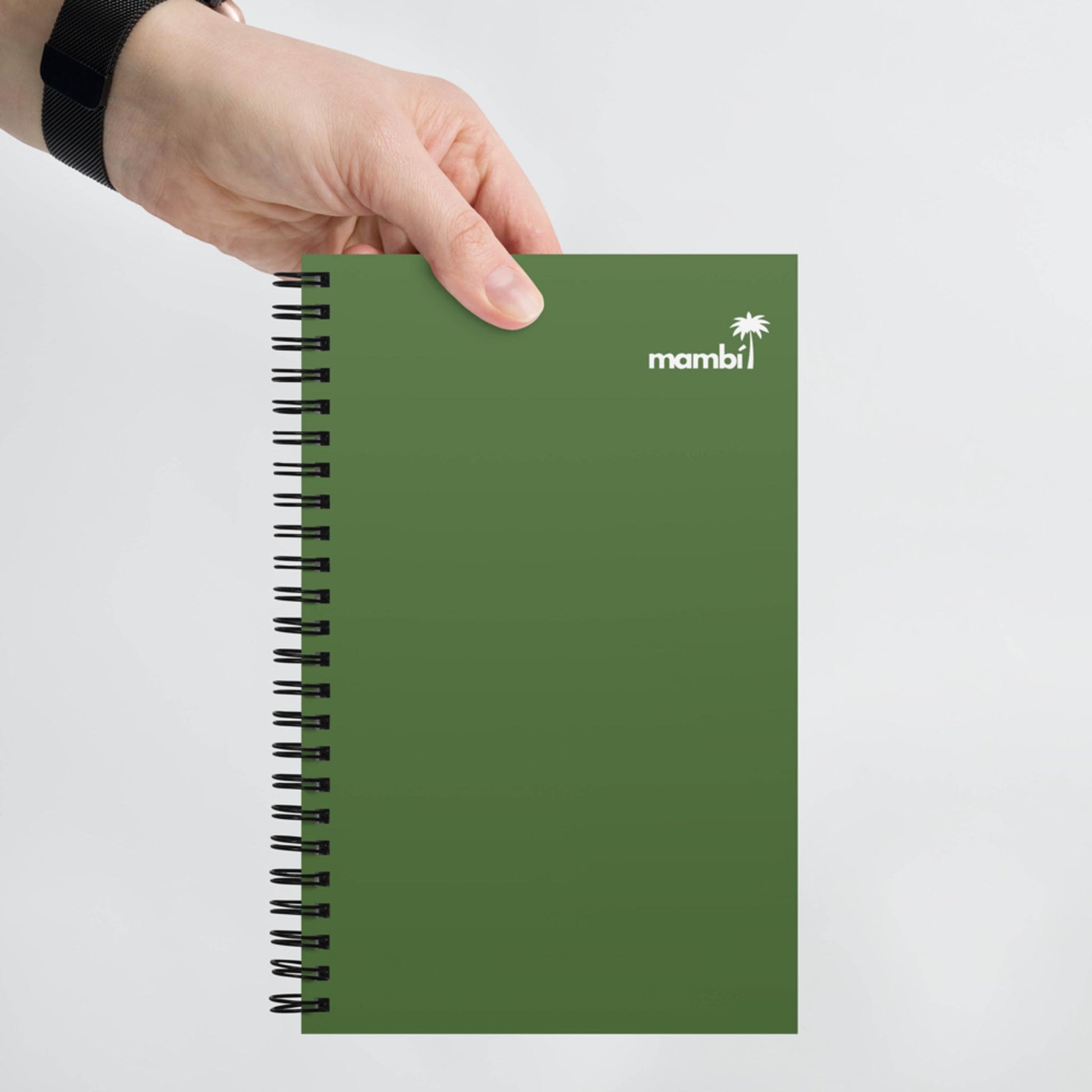 A hand holding a green, spiral notebook with mambi and a small palm tree logo printed in white on the top right corner of the cover.