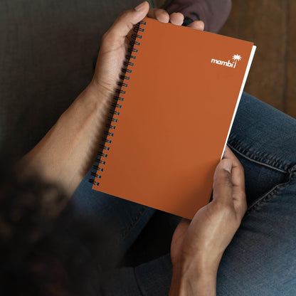 A person sits cross-legged, holding an orange wire-bound notebook with the word mambi and a small palm tree icon printed in white on the cover.