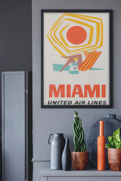 A framed vintage United Air Lines Miami travel poster featuring a beach scene and a touch of Mambí style hangs on a gray wall above a shelf adorned with vases, a potted plant, and a round black tray.