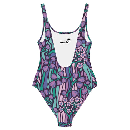 The Micaela One-Piece Swimsuit by Printful features a chic silhouette, scoop neckline, and vibrant floral print in purple, pink, teal, and green on stretchy fabric. The inner label displays “mambi” with a palm tree logo above it.