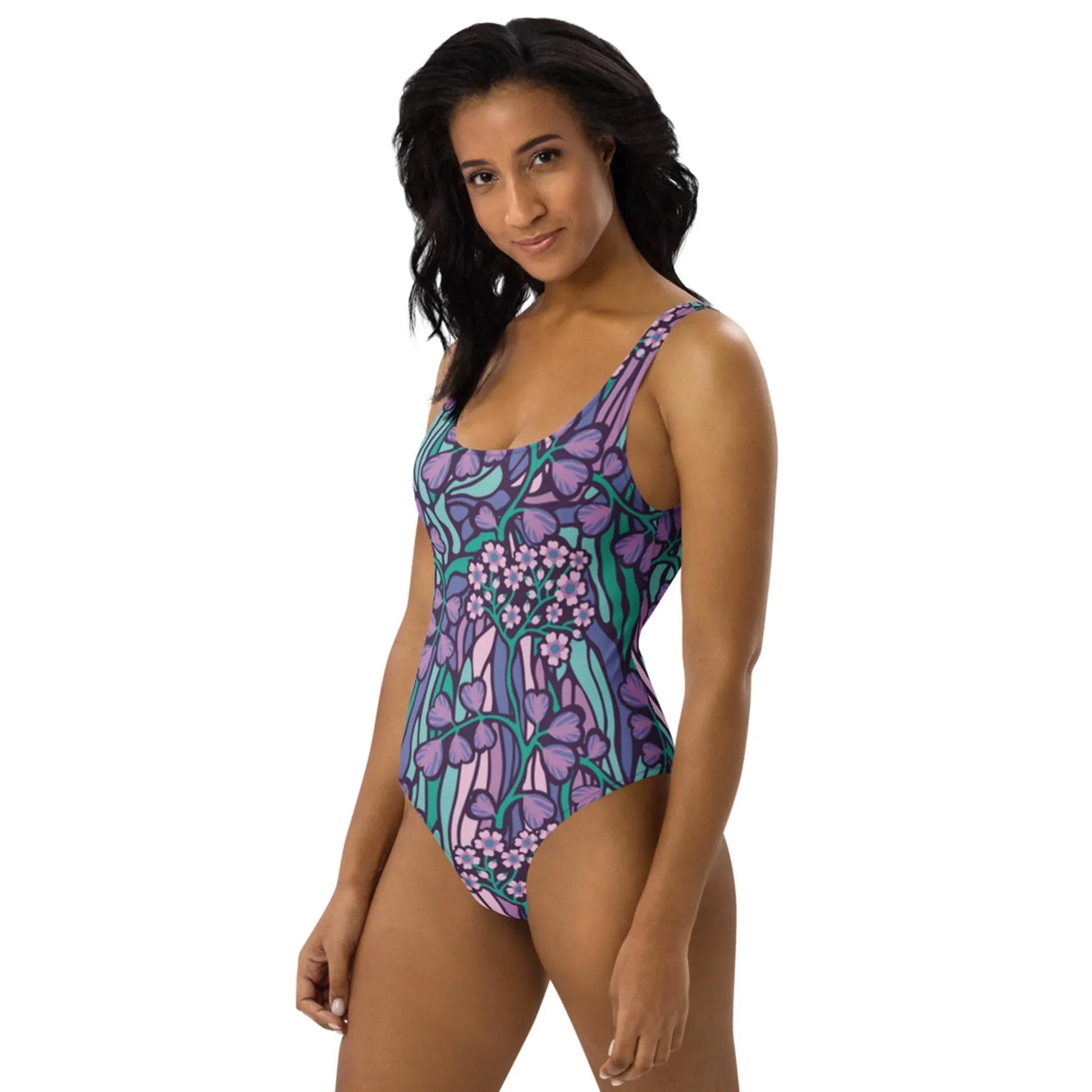 A woman with shoulder-length black hair wears the Micaela One-Piece Swimsuit by Printful in a chic purple and green floral design, standing against a white background and smiling slightly at the camera.