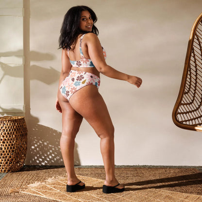 A woman stands on a woven rug wearing a colorful Mambí floral bikini and black sandals, looking over her shoulder and smiling. Sunlight casts shadows on the wall, with wicker baskets and a hanging chair nearby.