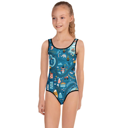 A young girl with long, blond hair in a ponytail is wearing a blue Mambí one-piece swimsuit featuring colorful, playful space-themed designs like rockets, stars, and planets. She stands with a slight smile.