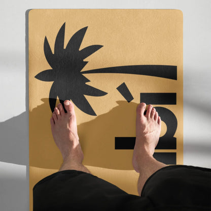 A pair of bare feet stand on a yellow Mambí mat featuring a black graphic of a bent palm tree and the number 5. The persons legs are partially visible, and shadows fall diagonally across the mat.