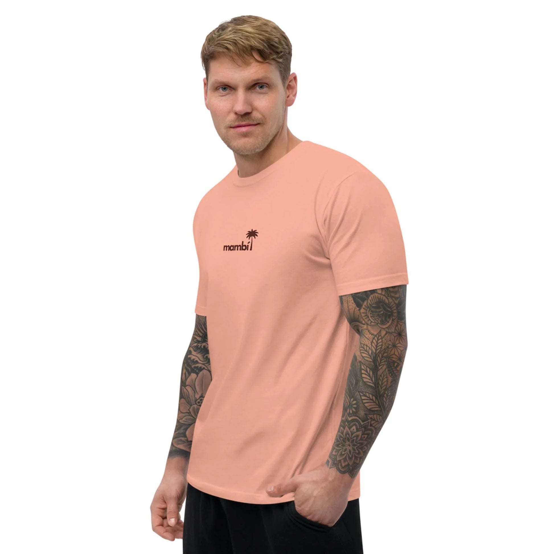 A tattooed man stands sideways with one hand in his pocket, wearing the Printful No Me Jodas Fitted Graphic Tee for men—a soft cotton peach shirt featuring mambi and a small palm tree graphic on the chest.