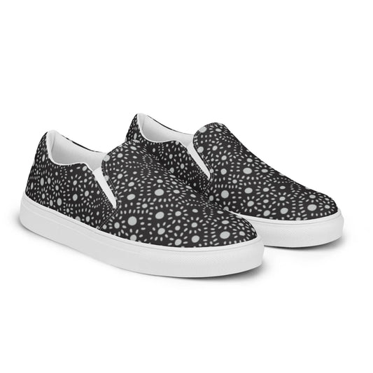 A pair of slip-on sneakers featuring a black background with a white geometric, kaleidoscopic dot pattern. The soles and inner lining are white, giving a modern, stylish Mambí-inspired appearance.